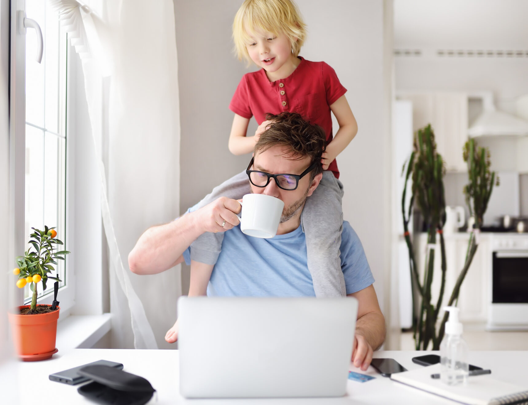 Man with his son on his shoulders, drinking coffee and doing work on his laptop.