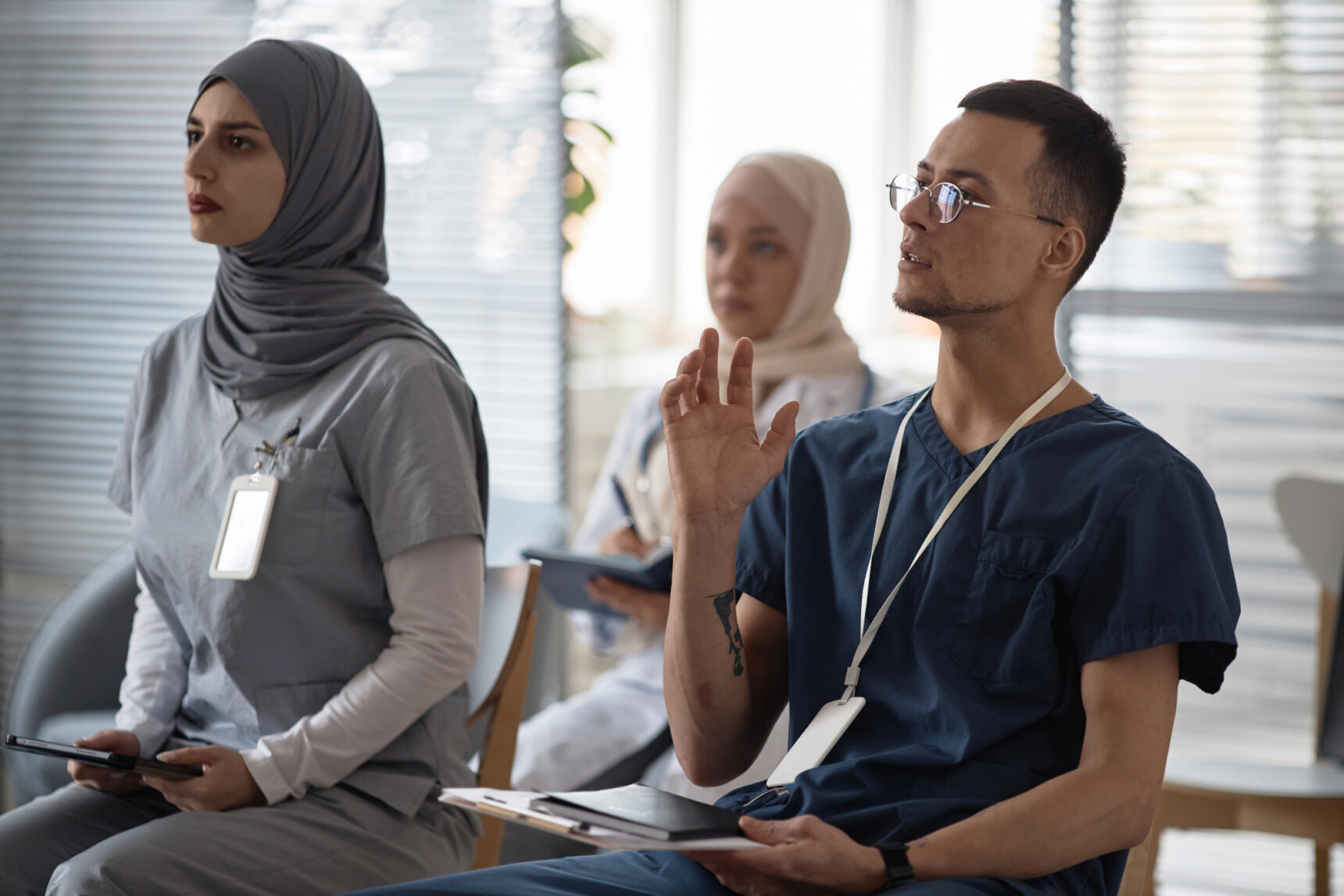 Nurse apprentices sat in scrubs with hand up asking a question.