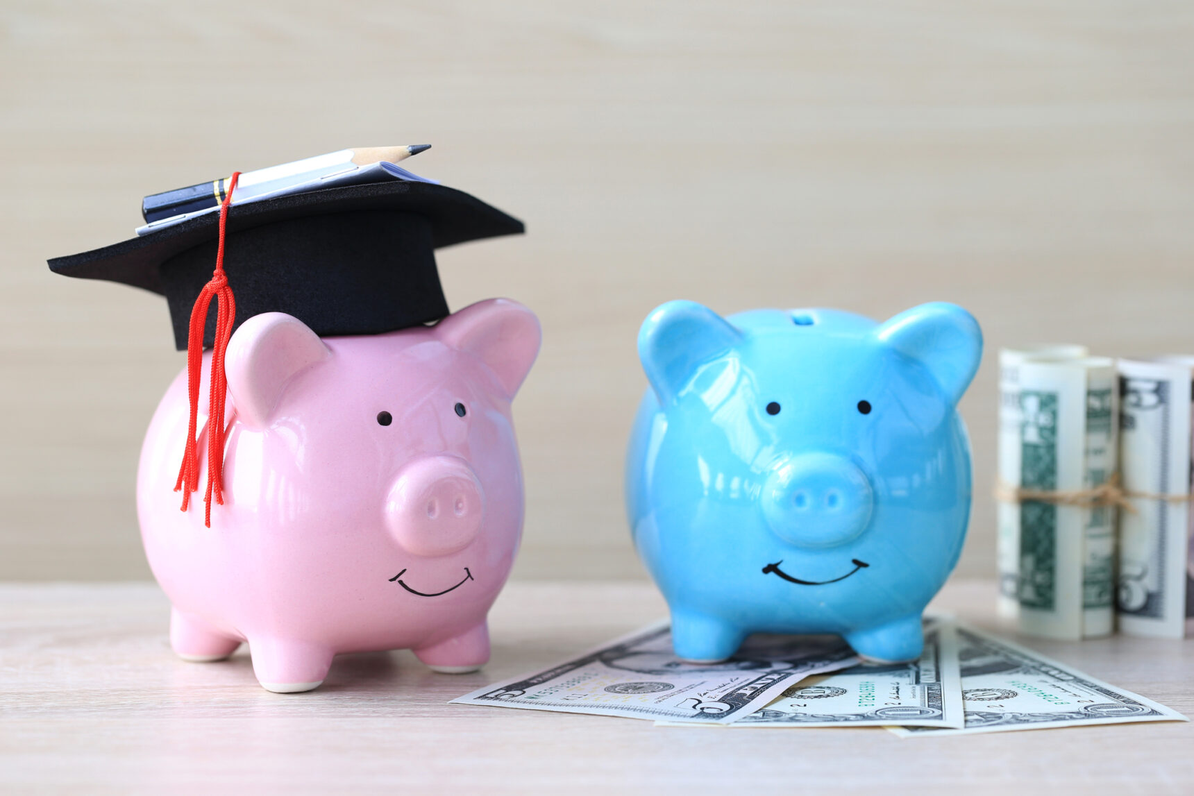 Graduation hat on pink piggy bank next to a blue piggy bank resting on banknotes