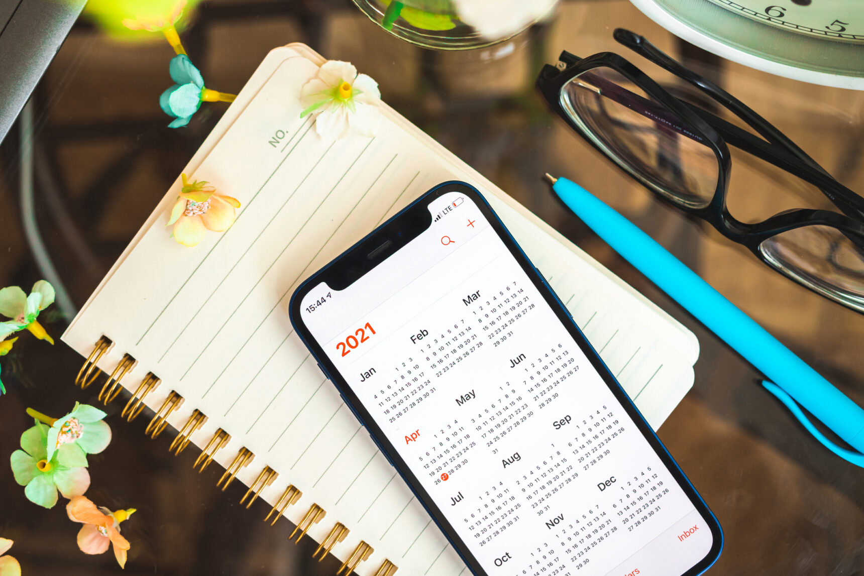 Phone with the calender app open, sat on top of a paper planner with a pen, glasses, and scattered flowers.