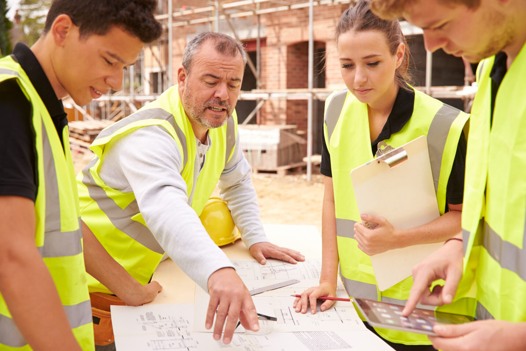 A teacher explains architect drawings to three student apprentices wearing fluorescent yellow hi-vis vests who are gathered on a building site.