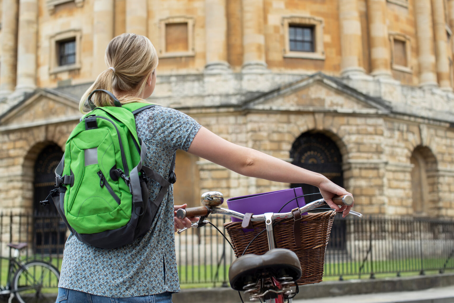 A student with a green backpack stands with her bicycle in front of Oxford University.